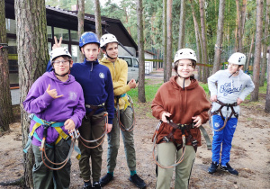 Grupa uczniów przygotowana do pokonania przeszkód w parku linowym ubrana w kaski oraz uprząż alpinistyczną.
